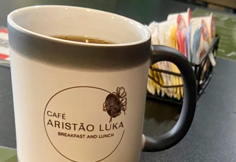 A mug of coffee branded with the Café Aristão Luka logo, served alongside a pastry